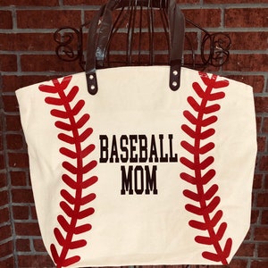 K&ouml;nnte beinhalten: Eine wei&szlig;e Canvas-Tragetasche mit schwarzen Henkeln und roten Baseballn&auml;hten. Die Tasche tr&auml;gt den Aufdruck "BASEBALL MOM" in Schwarz.
