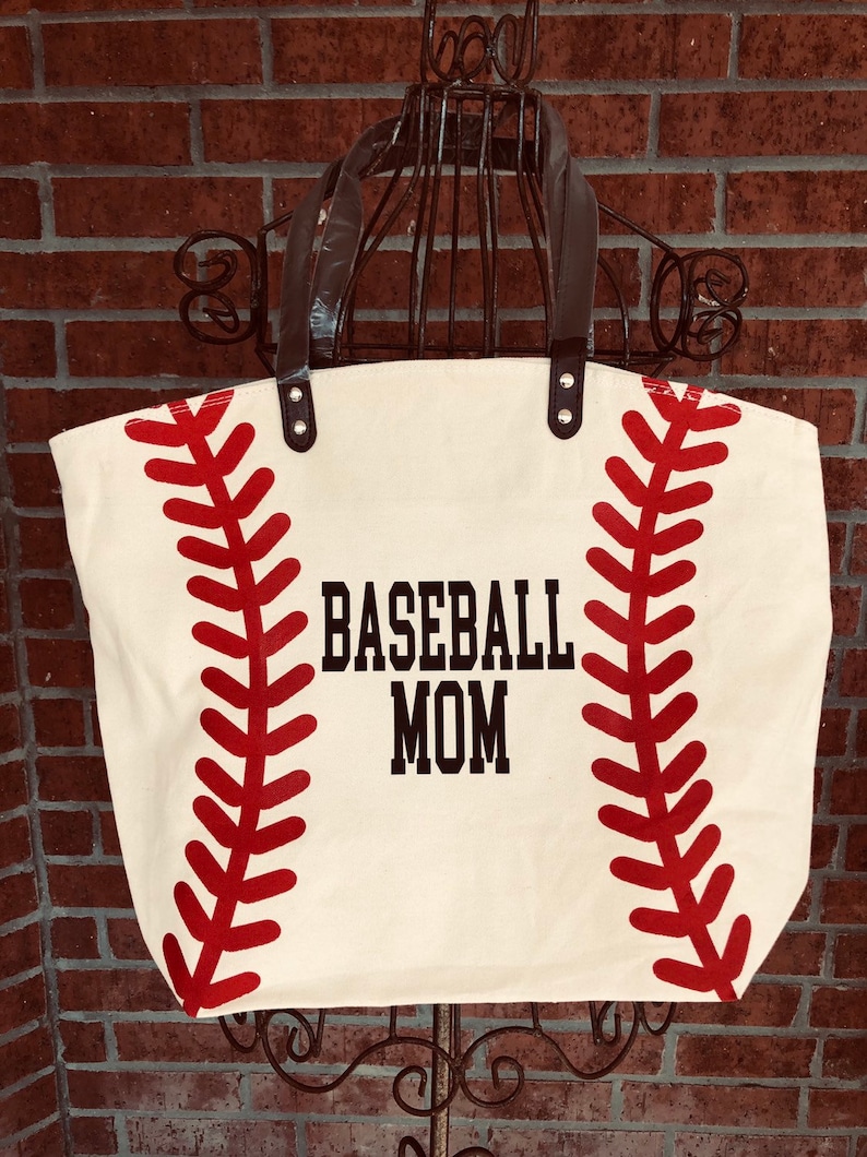 K&ouml;nnte beinhalten: Eine wei&szlig;e Canvas-Tragetasche mit schwarzen Henkeln und roten Baseballn&auml;hten. Die Tasche tr&auml;gt den Aufdruck "BASEBALL MOM" in Schwarz.