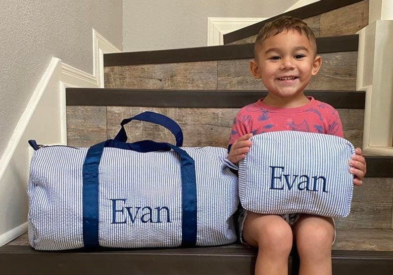 May include: A blue and white striped duffel bag and a matching toiletry bag with the name "Evan" embroidered on each. A young boy is sitting on a set of stairs holding the toiletry bag.