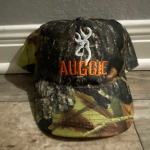 May include: Camouflage baseball cap with the name "AUGGIE" embroidered in orange. The cap features a white emblem and is designed with a brown, green, and yellow camouflage pattern.
