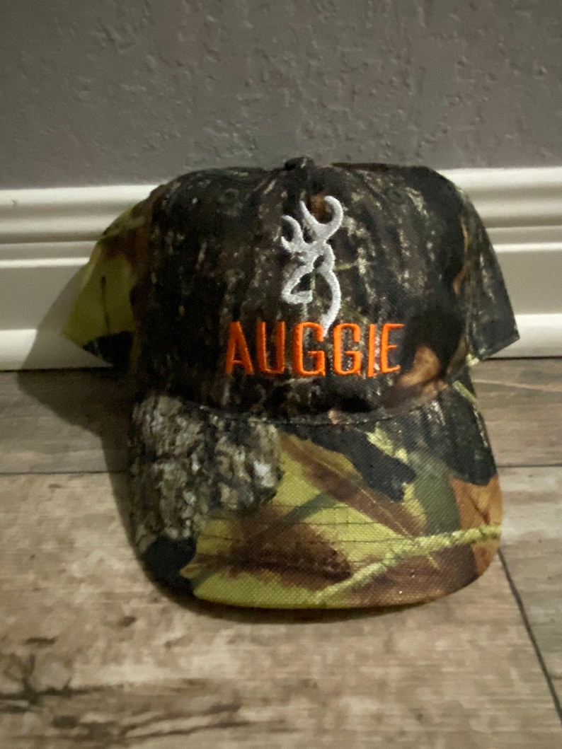 May include: Camouflage baseball cap with the name "AUGGIE" embroidered in orange. The cap features a white emblem and is designed with a brown, green, and yellow camouflage pattern.