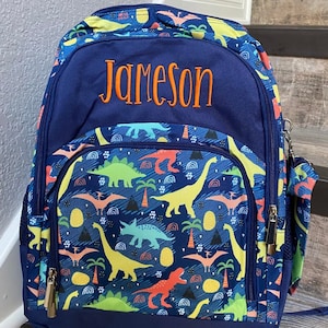 Może przedstawiać: Granatowy plecak z kolorowym wzorem dinozaur&oacute;w na przedniej kieszeni i bokach. Imię "Jameson" jest wyhaftowane na pomarańczowo na g&oacute;rze. Posiada boczną kieszeń, zapięcia na zamek błyskawiczny i regulowane paski.