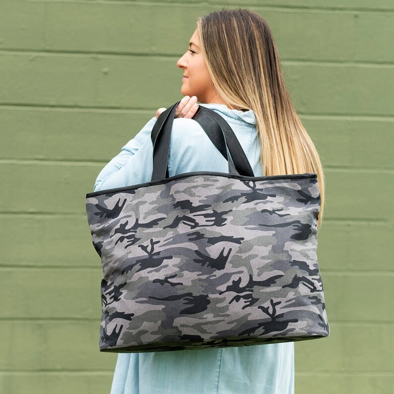 May include: A large black and gray camouflage tote bag with black handles. The bag is made of canvas and has a zipper closure.