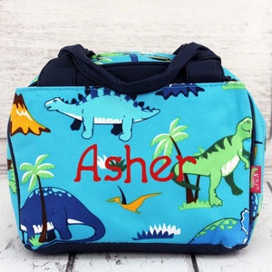 May include: A blue canvas lunch bag with a dinosaur print and the name "Asher" embroidered in red. The bag has two navy blue handles.