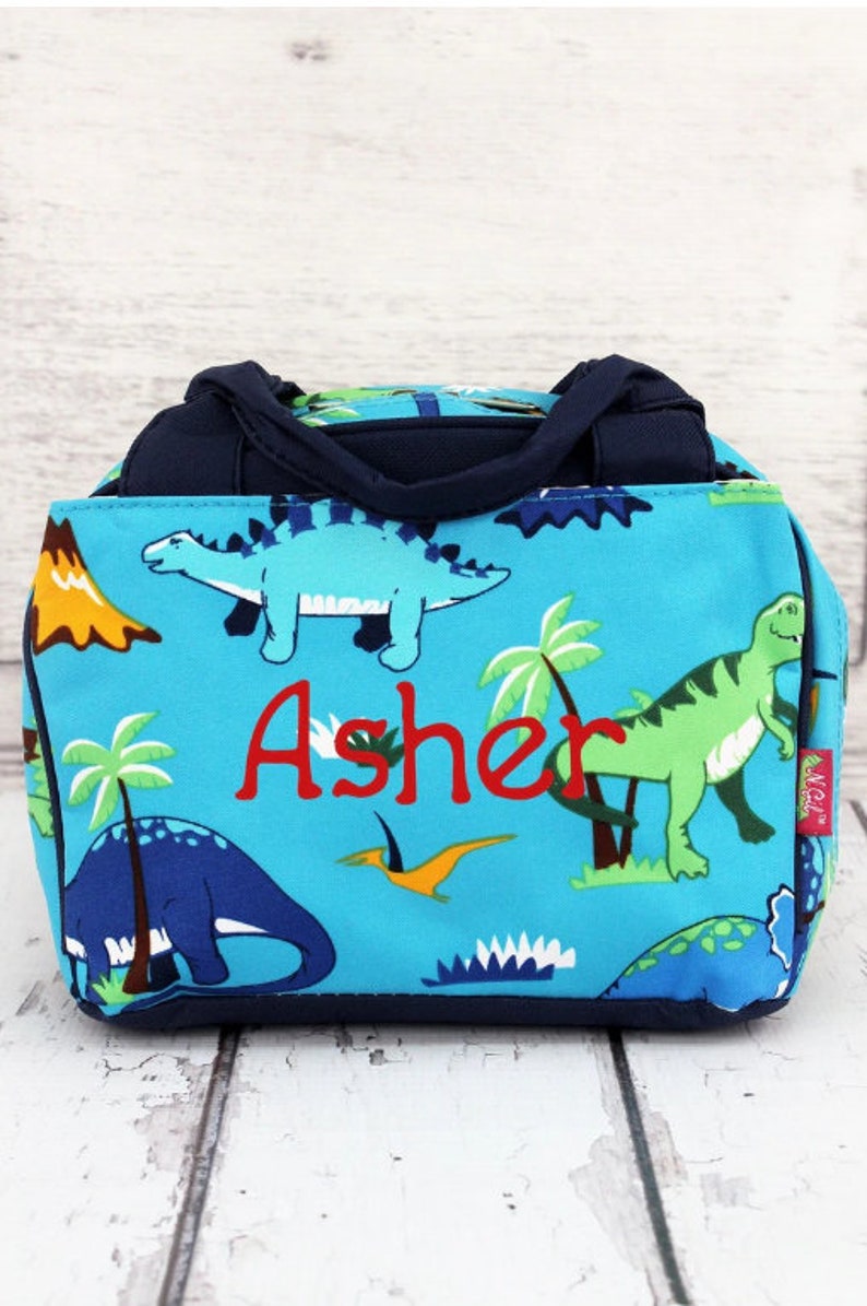 May include: A blue canvas lunch bag with a dinosaur print and the name "Asher" embroidered in red. The bag has two navy blue handles.