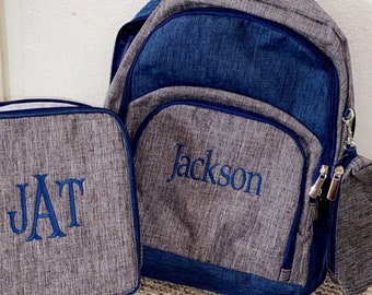 May include: A gray and navy blue backpack and matching lunch bag set. The backpack features the name Jackson embroidered in blue thread. The lunch bag features the monogram JAT embroidered in blue thread. Both items have a textured fabric finish.