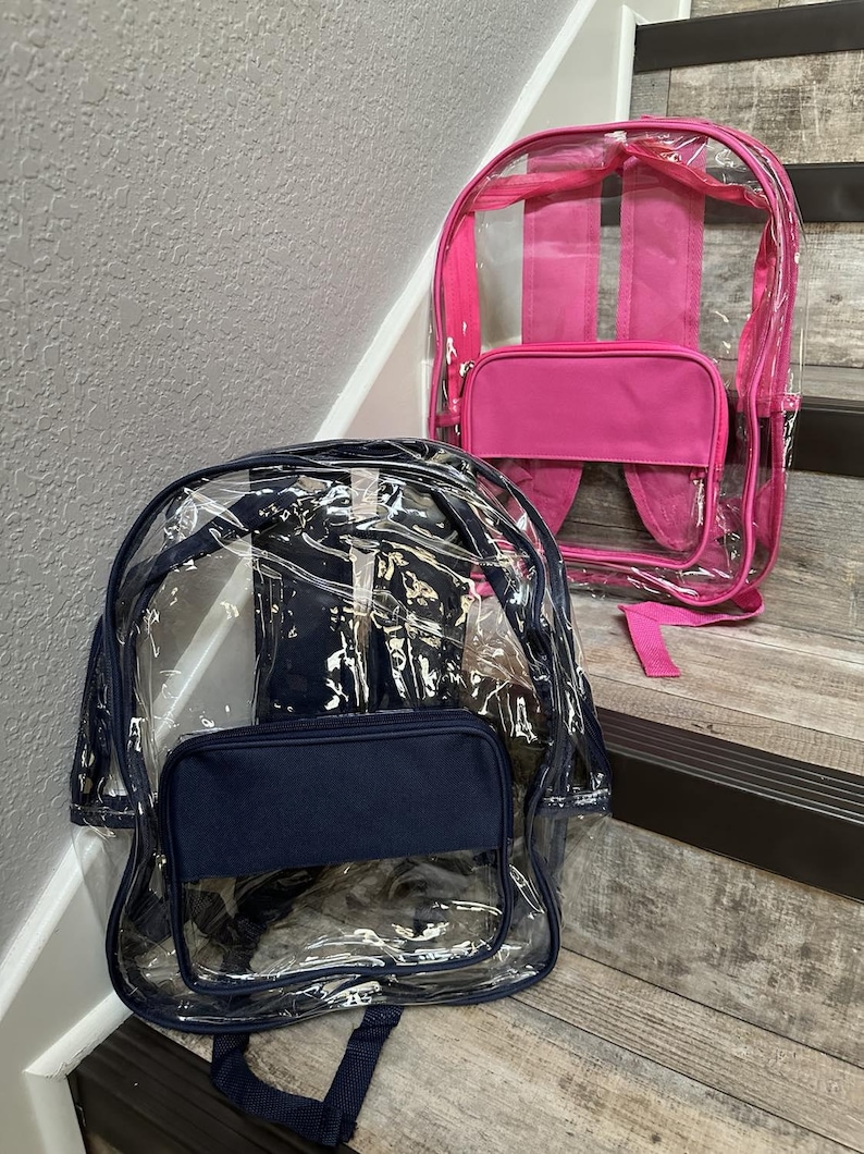 May include: Two clear backpacks with a front zippered pocket. One backpack has a pink pocket and the other has a navy blue pocket.