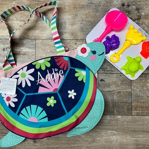 May include: A pink and blue beach bag with a turtle design and the name "Addie" embroidered on it. The bag comes with a set of colorful plastic beach toys, including a shovel, a crown, a seashell, and a turtle.