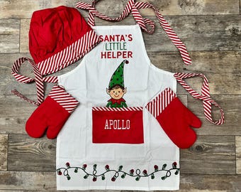 Könnte beinhalten: Weiße Weihnachts-Schürze mit rot-weißem Streifenbesatz. Die Schürze hat eine Tasche mit dem Namen "Apollo" bestickt. Zur Schürze gehören auch eine passende Kochmütze und zwei Topflappen. Auf der Schürze steht "Santa's Little Helper".