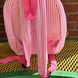 May include: Pink and white striped backpack with adjustable pink straps. The backpack has a rounded shape and features a small pocket on each side. The straps are attached with black plastic buckles. The backpack is made of a textured fabric.