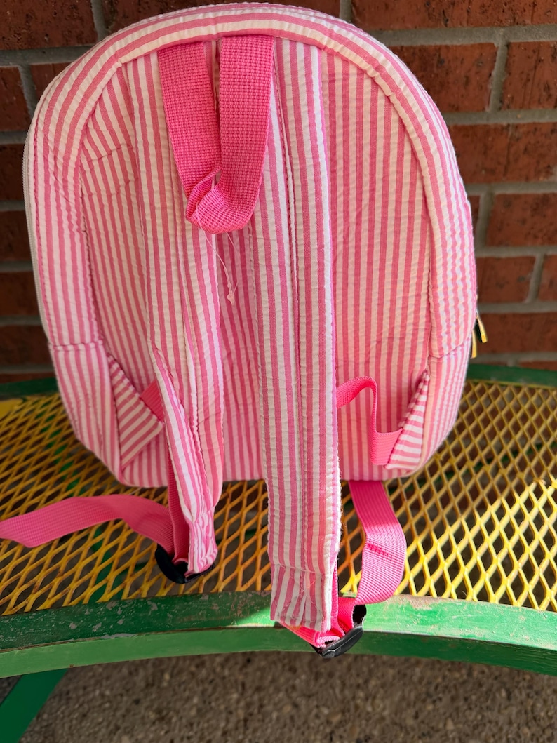 May include: Pink and white striped backpack with adjustable pink straps. The backpack has a rounded shape and features a small pocket on each side. The straps are attached with black plastic buckles. The backpack is made of a textured fabric.