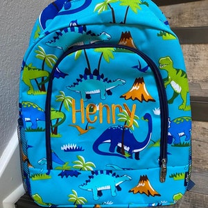 May include: A blue backpack with a dinosaur pattern. The backpack has a mesh side pocket and a zipped front pocket. The name "Henry" is embroidered on the front of the backpack.