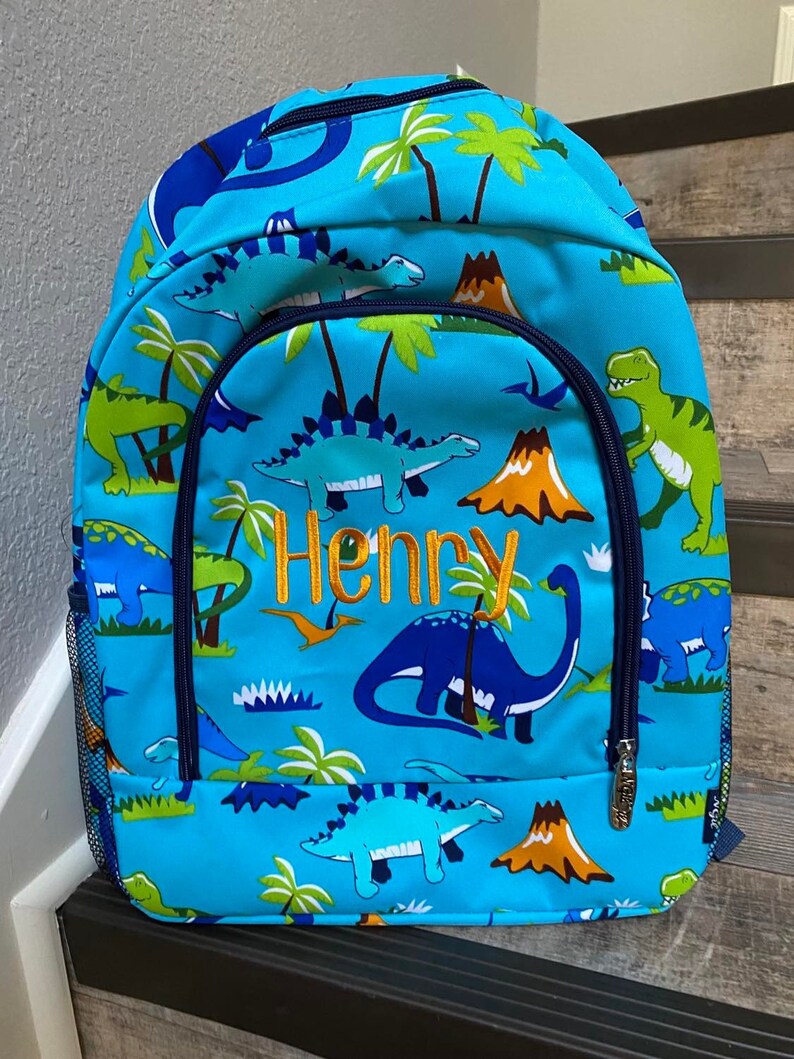 May include: A blue backpack with a dinosaur pattern. The backpack has a mesh side pocket and a zipped front pocket. The name "Henry" is embroidered on the front of the backpack.