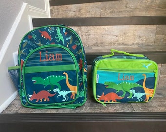 May include: A green and blue dinosaur-themed backpack and lunch bag set with the name "Liam" embroidered on both. The backpack has a mesh side pocket and a green zipper pull. The lunch bag has a green handle.
