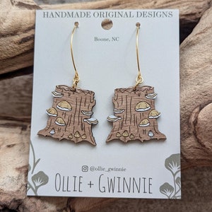 May include: A pair of wooden earrings featuring a mushroom design. The earrings are brown with yellow mushrooms and are hanging from gold hooks. The earrings are displayed on a white card with the text "HANDMADE ORIGINAL DESIGNS" and "Boone, NC". The card also includes the text "@ollie_gwinnie" and "OLLIE + GWINNIE".