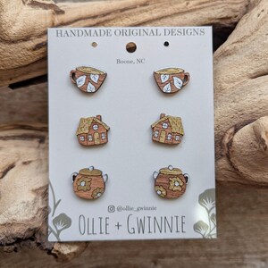 May include: Wooden stud earrings in the shape of a teacup, house, and teapot. The earrings are brown with white and yellow accents. The card reads "HANDMADE ORIGINAL DESIGNS" and "OLLIE + GWINNIE".