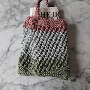May include: A multi-colored crocheted string bag with a newspaper inside. The bag is made of three colors: pink, gray, and green.