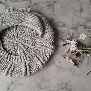 K&ouml;nnte beinhalten: Eine graue Strickm&uuml;tze mit einem gesprenkelten Muster. Die M&uuml;tze ist zur H&auml;lfte gefaltet und liegt flach auf einer Marmorfl&auml;che.