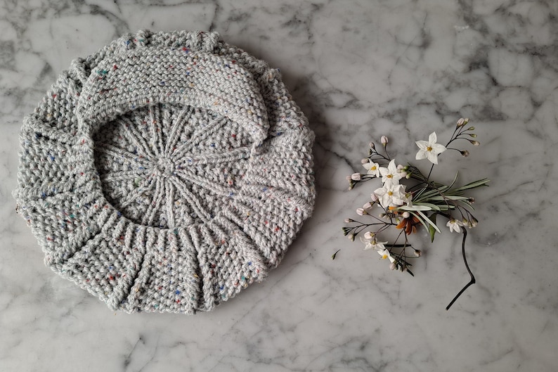 K&ouml;nnte beinhalten: Eine graue Strickm&uuml;tze mit einem gesprenkelten Muster. Die M&uuml;tze ist zur H&auml;lfte gefaltet und liegt flach auf einer Marmorfl&auml;che.