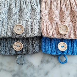 Puede incluir: Tres gorros de punto con botones de madera que dicen "Handmade" en forma de corazón. Los gorros son grises, rosa y azul.