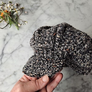 K&ouml;nnte beinhalten: Eine schwarze und wei&szlig;e Tweed-Newsboy-M&uuml;tze mit braunen Sprenkeln. Die M&uuml;tze ist aus weichem, gestricktem Stoff gefertigt und hat ein klassisches, zeitloses Design.