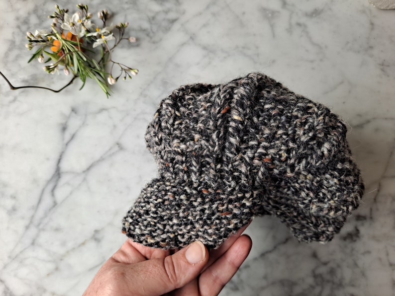 K&ouml;nnte beinhalten: Eine schwarze und wei&szlig;e Tweed-Newsboy-M&uuml;tze mit braunen Sprenkeln. Die M&uuml;tze ist aus weichem, gestricktem Stoff gefertigt und hat ein klassisches, zeitloses Design.