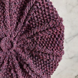 K&ouml;nnte beinhalten: Nahaufnahme einer dunkelvioletten Strickm&uuml;tze mit einem strukturierten Muster. Die M&uuml;tze ist aus weichem Garn gefertigt und hat ein einzigartiges Design.