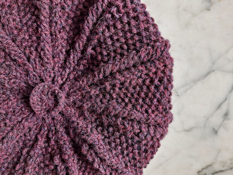 K&ouml;nnte beinhalten: Nahaufnahme einer dunkelvioletten Strickm&uuml;tze mit einem strukturierten Muster. Die M&uuml;tze ist aus weichem Garn gefertigt und hat ein einzigartiges Design.