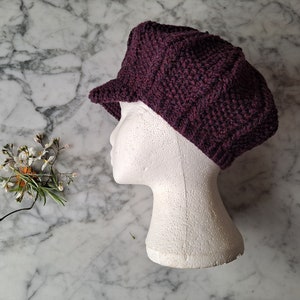 K&ouml;nnte beinhalten: Eine dunkelviolette Strickm&uuml;tze im Newsboy-Stil mit leicht eingefallener Krone und kurzem Schirm. Die M&uuml;tze wird auf einem wei&szlig;en Mannequin-Kopf pr&auml;sentiert.