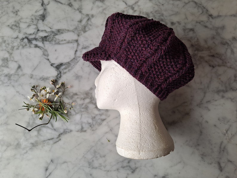 K&ouml;nnte beinhalten: Eine dunkelviolette Strickm&uuml;tze im Newsboy-Stil mit leicht eingefallener Krone und kurzem Schirm. Die M&uuml;tze wird auf einem wei&szlig;en Mannequin-Kopf pr&auml;sentiert.