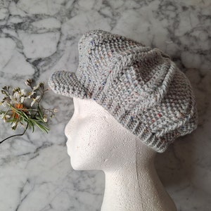 Puede incluir: Una gorra de newsboy de punto gris con un patrón de motas multicolores. La gorra está en una cabeza de maniquí blanca.