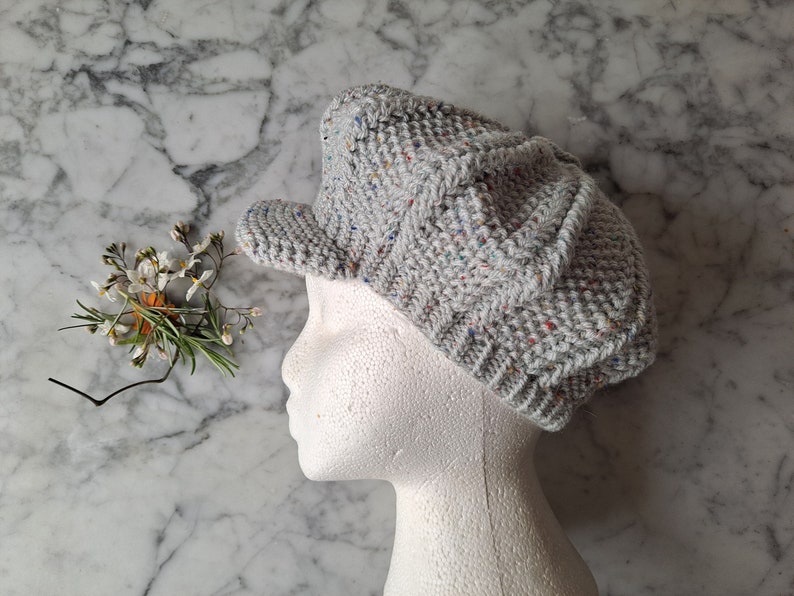 K&ouml;nnte beinhalten: Eine graue Strickm&uuml;tze im Newsboy-Stil mit einem mehrfarbigen Fleckenmuster. Die M&uuml;tze sitzt auf einem wei&szlig;en Mannequin-Kopf.