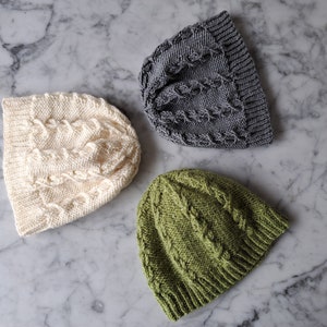 May include: Three knitted hats in different colors: cream, gray, and green. Each hat has a cable knit pattern.