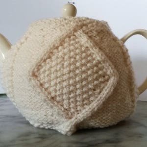 May include: A white knitted teapot cozy with a diamond pattern. The cozy is made of a soft, textured yarn and is designed to keep tea warm.