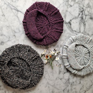 K&ouml;nnte beinhalten: Drei gestrickte Baskenm&uuml;tzen in verschiedenen Farben: dunkelgrau mit wei&szlig;en Flecken, hellgrau mit wei&szlig;en Flecken und ein tiefes Lila. Die Baskenm&uuml;tzen sind in Dreiecksform auf einer wei&szlig;en Marmorfl&auml;che mit einem kleinen Strau&szlig; wei&szlig;er Blumen in der Mitte angeordnet.