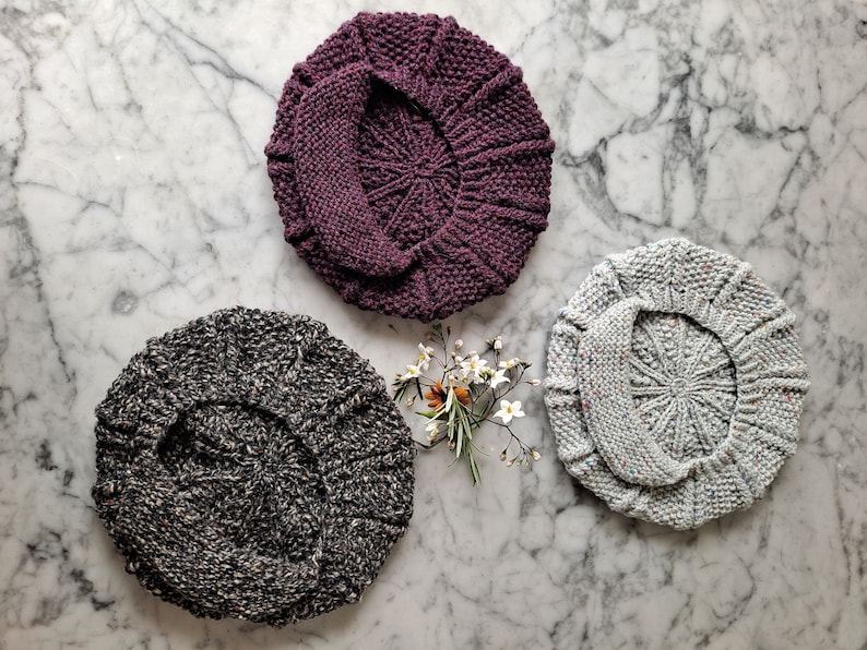 K&ouml;nnte beinhalten: Drei gestrickte Baskenm&uuml;tzen in verschiedenen Farben: dunkelgrau mit wei&szlig;en Flecken, hellgrau mit wei&szlig;en Flecken und ein tiefes Lila. Die Baskenm&uuml;tzen sind in Dreiecksform auf einer wei&szlig;en Marmorfl&auml;che mit einem kleinen Strau&szlig; wei&szlig;er Blumen in der Mitte angeordnet.