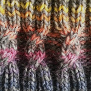 May include: Close-up of a knitted fabric with a multi-colored striped pattern. The fabric is made with a variety of shades of gray, pink, orange, and yellow yarn.