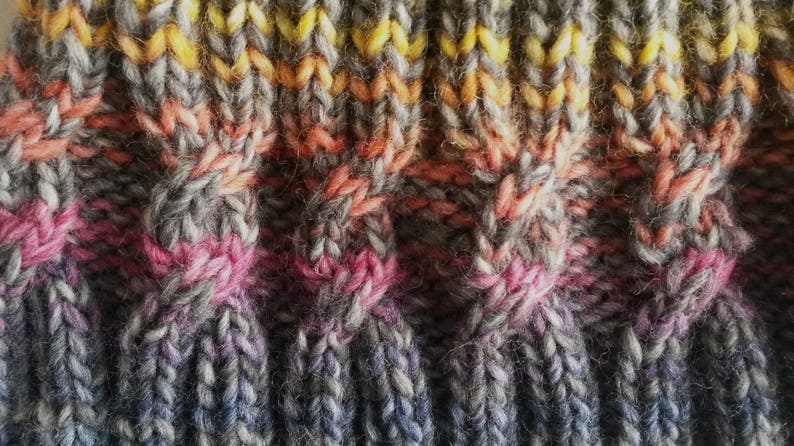 May include: Close-up of a knitted fabric with a multi-colored striped pattern. The fabric is made with a variety of shades of gray, pink, orange, and yellow yarn.