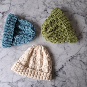May include: Three knitted hats in different colors: light blue, green, and cream. Each hat has a cable knit pattern.