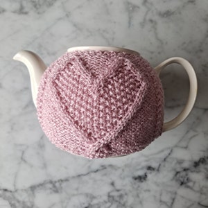 Puede incluir: Una tetera blanca con un calienta-teteras de punto rosa en forma de corazón. El calienta-teteras está hecho de un hilo suave y texturizado.