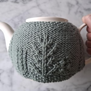 May include: A white ceramic teapot with a sage green knitted tea cozy. The cozy features a leaf pattern. The teapot is being held by a person.