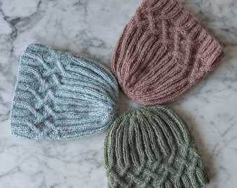 May include: Three knitted hats in different colours: light blue, dusty rose, and olive green. The hats have a cable knit pattern and are arranged on a white marble surface.