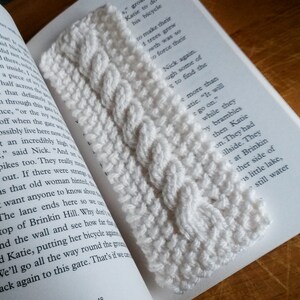 May include: A white crocheted bookmark with a cable stitch pattern. The bookmark is lying on a book with text.
