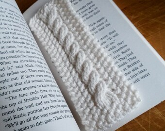 May include: A white crocheted bookmark with a cable stitch pattern. The bookmark is lying on a book with text.