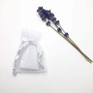 May include: A white organza gift bag with a silver ribbon closure, containing a small, square item. Dried lavender sprigs with purple flowers are arranged next to the bag on a white background. The image is a top-down view.