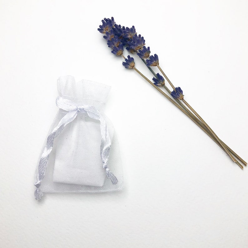 May include: A white organza gift bag with a silver ribbon closure, containing a small, square item. Dried lavender sprigs with purple flowers are arranged next to the bag on a white background. The image is a top-down view.
