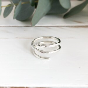 May include: A silver spiral ring with a hammered finish. The ring features two loops and an open end. The ring is displayed on a white surface, with green foliage in the background.