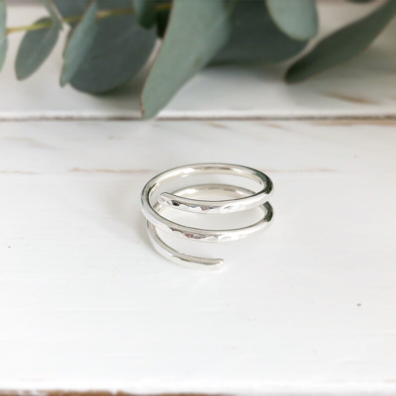 May include: A silver spiral ring with a hammered finish. The ring features two loops and an open end. The ring is displayed on a white surface, with green foliage in the background.