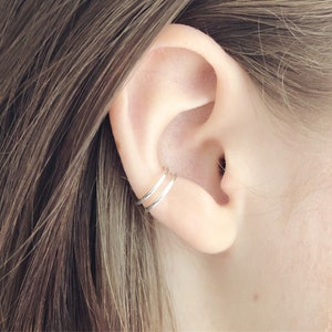 May include: A close-up of a person's ear with two silver ear cuffs. The cuffs are thin and delicate, and they are worn on the upper part of the ear.