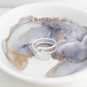 May include: A silver spiral ring with a hammered texture. The ring sits on a white and grey marbled ceramic dish with gold accents. The ring's design features multiple loops, creating a unique and modern look.